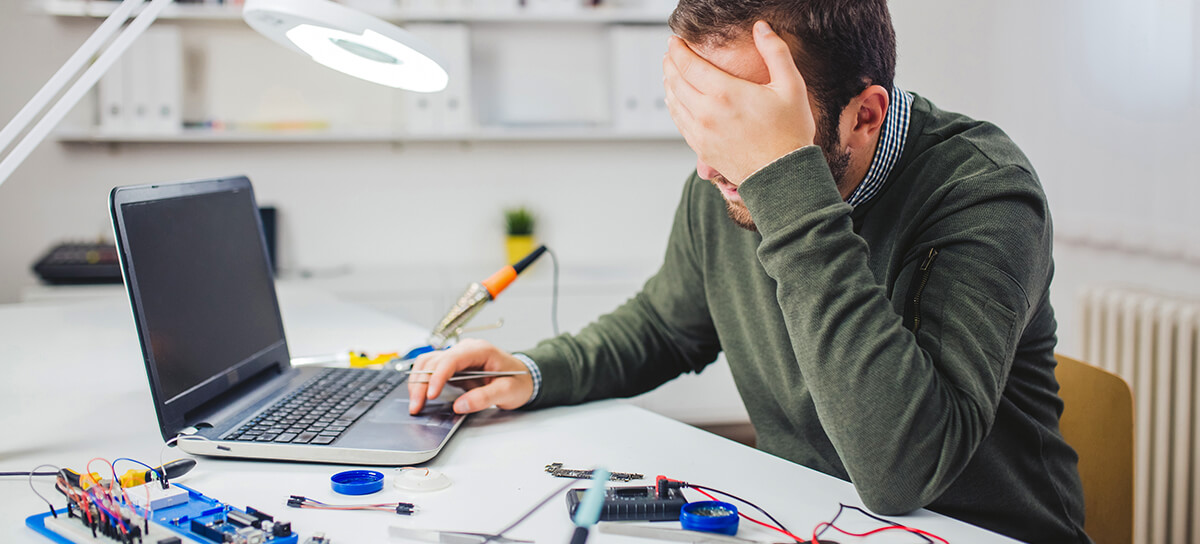 MainImage_1200x544 A tired man sitting at a table with a laptop, dev kit, and tools on it.