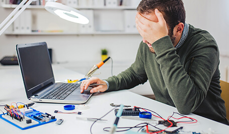 A tired man sitting at a table with a laptop, dev kit, and tools on it.