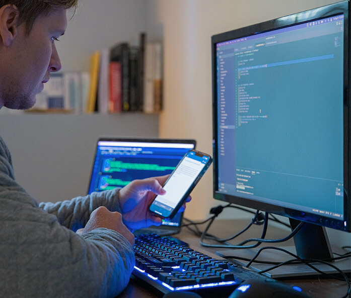A programmer uses a laptop, computer, and smartphone to work on code for a custom software development project.