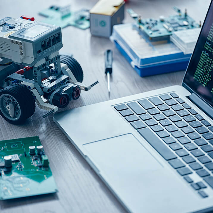 A small robot, a PCB, and a laptop on a desk