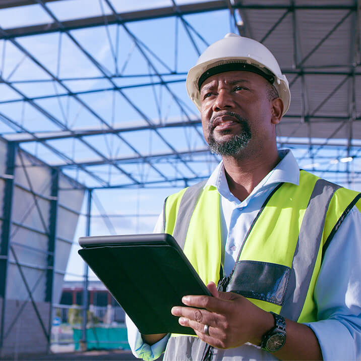 A man in a fluorescent vest and helmet holds a tablet in his hands.